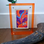 A photo of a colorful, illustrated dragon with a pink bow on its head holding a white fluffy cat with an acrylic orange frame. There is a small green dragon toy placed on top of the frame and a carabao horn placed in front of the art in the foreground.