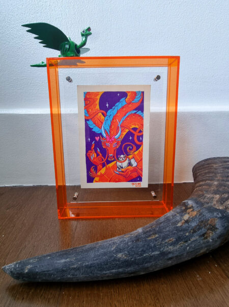 A photo of a colorful, illustrated dragon with a pink bow on its head holding a white fluffy cat with an acrylic orange frame. There is a small green dragon toy placed on top of the frame and a carabao horn placed in front of the art in the foreground.