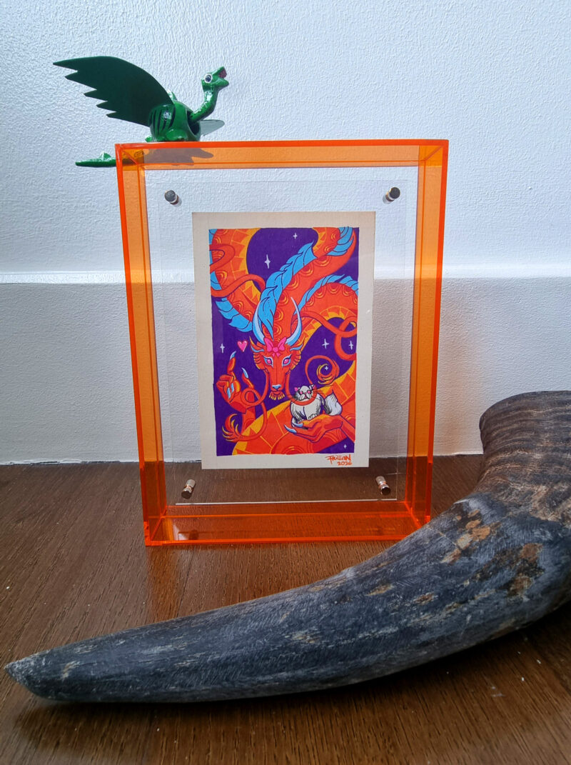A photo of a colorful, illustrated dragon with a pink bow on its head holding a white fluffy cat with an acrylic orange frame. There is a small green dragon toy placed on top of the frame and a carabao horn placed in front of the art in the foreground.