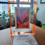 A photo of a colorful, illustrated dragon with a pink bow on its head holding a white fluffy cat with an acrylic orange frame placed on top of a wood table. There is a certificate of authenticity as a piece of small paper placed in front of the frame. An apartment window and potted plants are seen in the background.