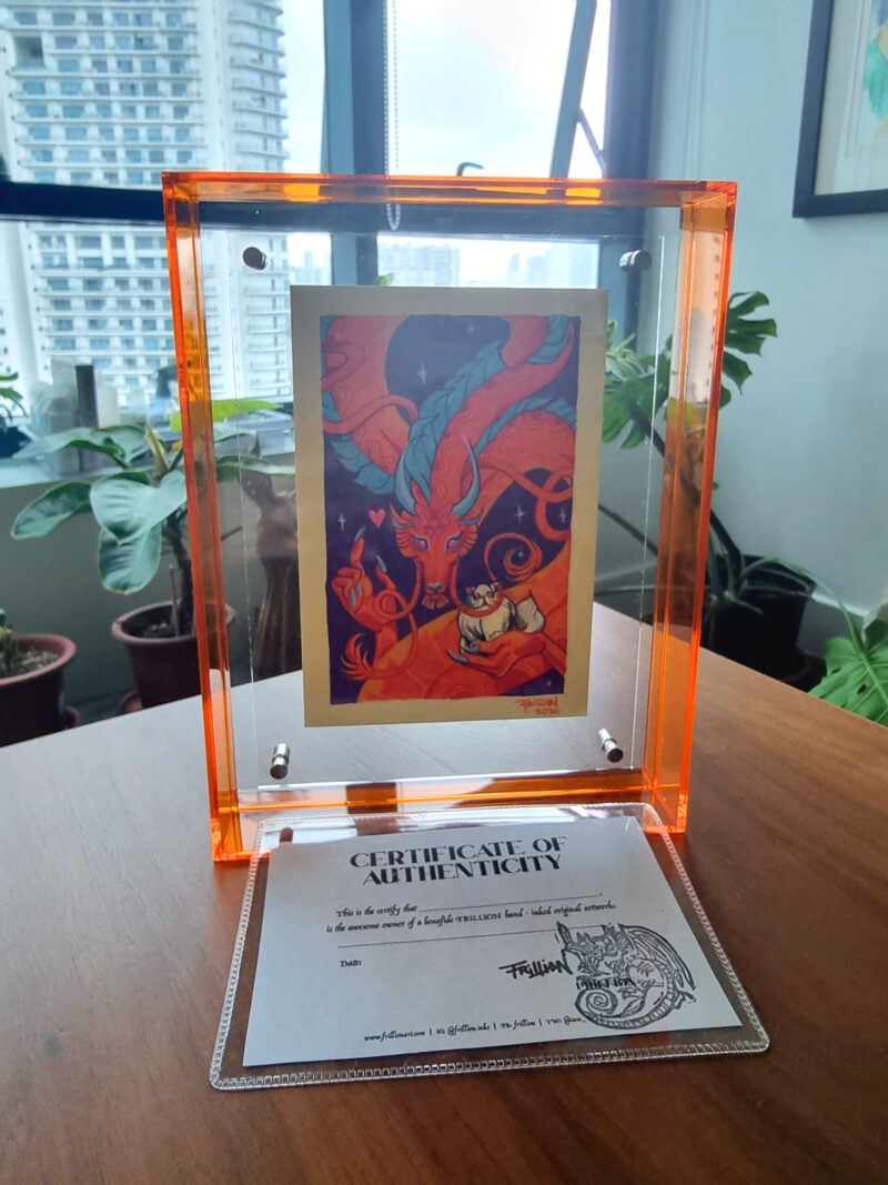 A photo of a colorful, illustrated dragon with a pink bow on its head holding a white fluffy cat with an acrylic orange frame placed on top of a wood table. There is a certificate of authenticity as a piece of small paper placed in front of the frame. An apartment window and potted plants are seen in the background.