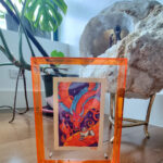 A photo of a colorful, illustrated dragon with a pink bow on its head holding a white fluffy cat with an acrylic orange frame. There is a plant and decorative stone in the background.