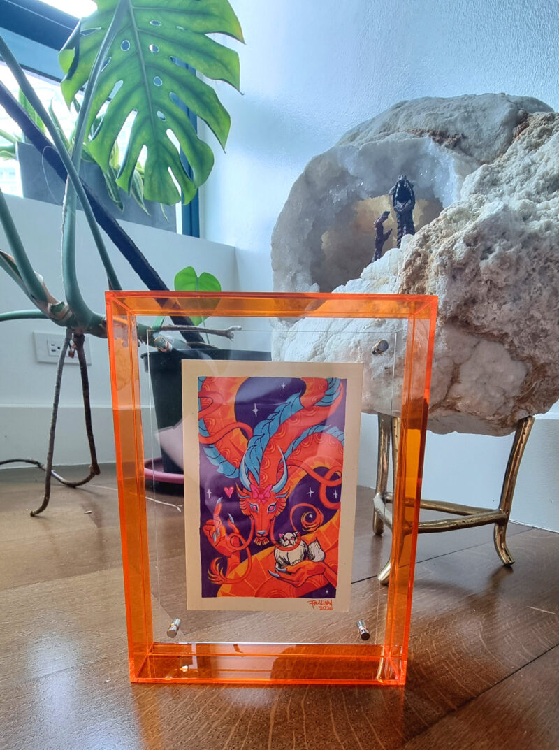 A photo of a colorful, illustrated dragon with a pink bow on its head holding a white fluffy cat with an acrylic orange frame. There is a plant and decorative stone in the background.