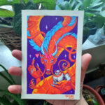 A photo of a colorful, illustrated dragon with a pink bow on its head holding a white fluffy cat. It is held by a right hand with potted plants in the background.
