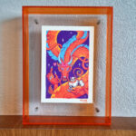 A photo of a colorful, illustrated dragon with a pink bow on its head holding a white fluffy cat with an acrylic orange frame. It is placed on top of a wooden ledge and in front of a concrete background.