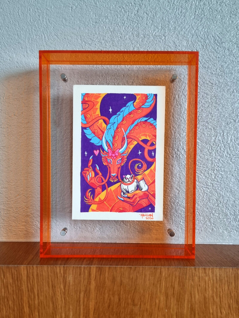 A photo of a colorful, illustrated dragon with a pink bow on its head holding a white fluffy cat with an acrylic orange frame. It is placed on top of a wooden ledge and in front of a concrete background.