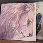photo of a painting with jars in the background. Square watercolor painting is of a lion head in pale violets, pinks and creams