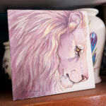 photo of a painting with jars in the background. Square watercolor painting is of a lion head in pale violets, pinks and creams