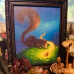 Framed squirrel painting in studio surrounded by wooden mushroom and artificial flowers on desk.