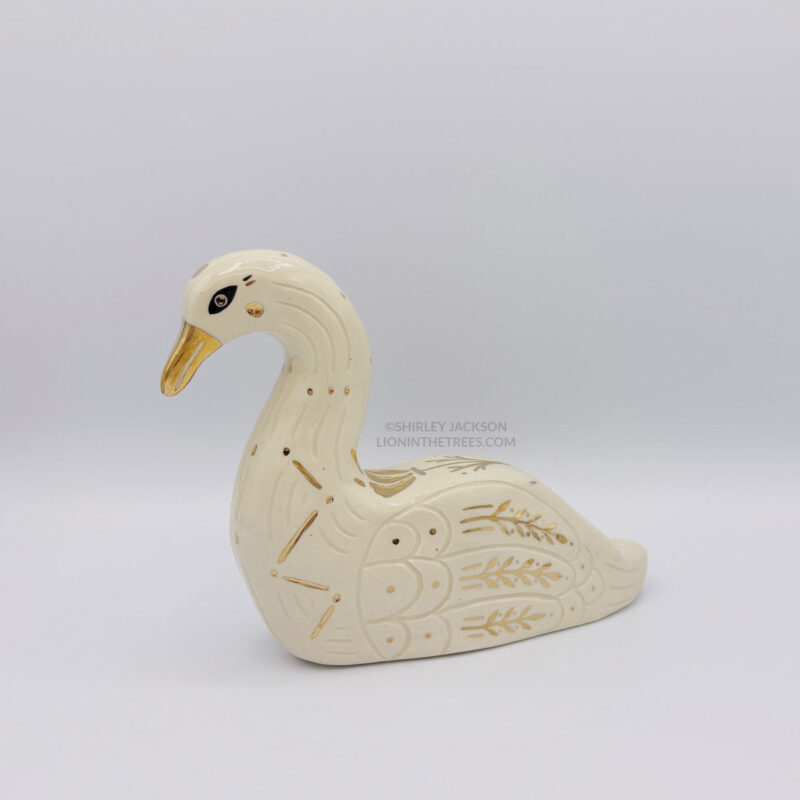 A ceramic swan sculpture with sgraffito details done with white clay and gold luster details such as a golden beak, a constellation that runs on the side of it's chest, and floral details on the wings. This side shows a golden eye.
